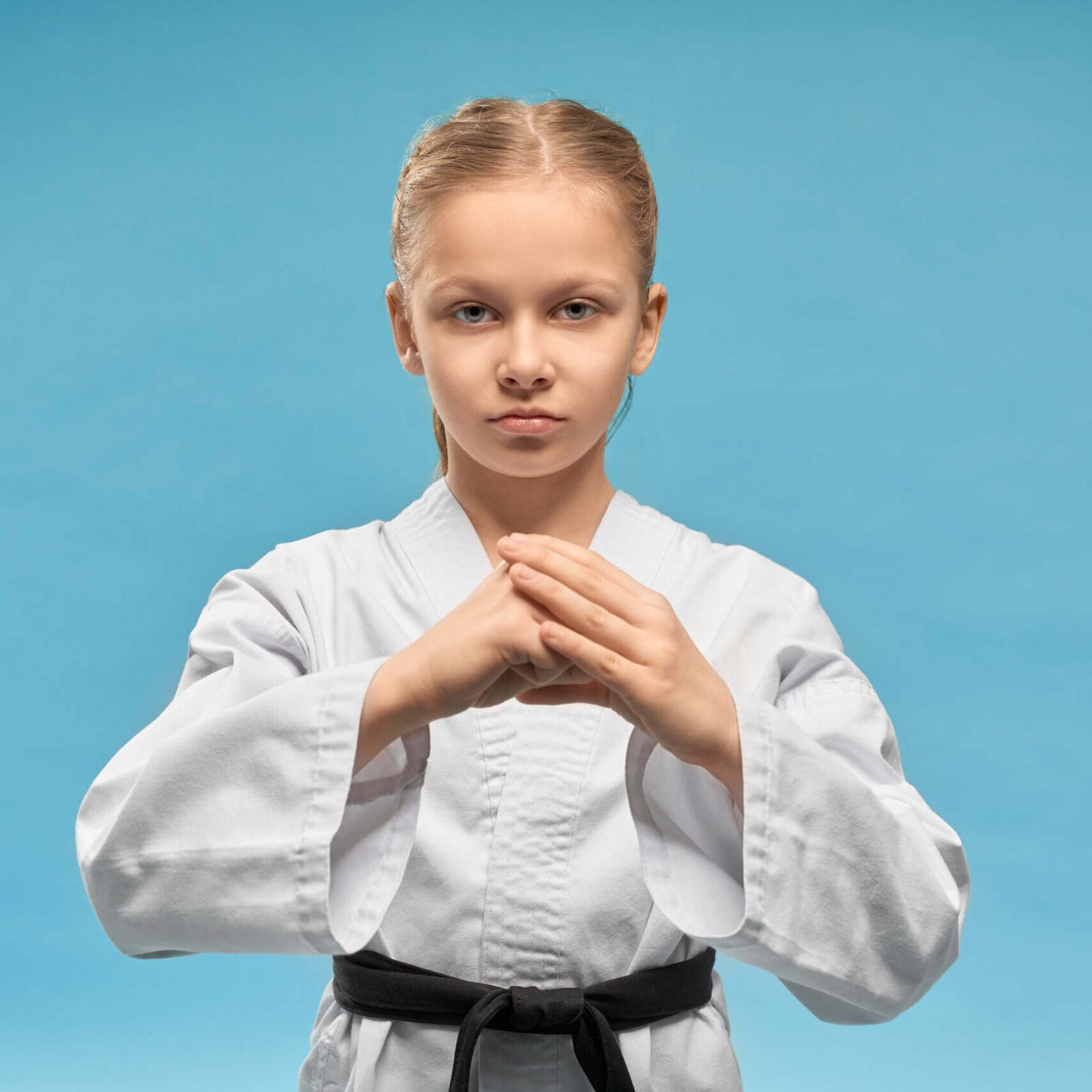 Young concentrated karate girl practicing hands position. Pretty junior in white kimono with black belt showing fist. Confident child posing on blue background, looking at camera.